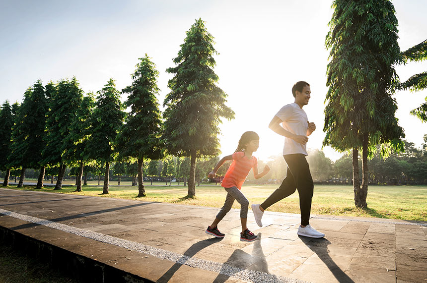 Father and daughter jogging together along a tree-lined park path.