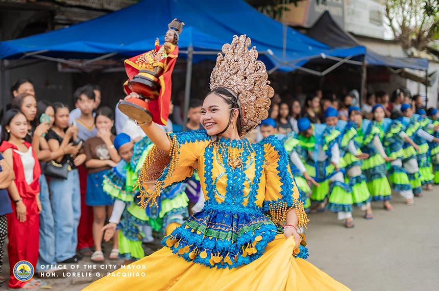 Street dancer in a gold headdress and yellow-blue costume leading performers at a Kadayawan Festival street dance