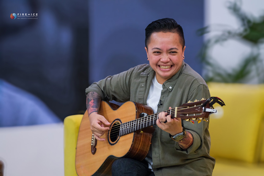 A photo of musician Ice Seguerra smiling as he plays his guitar while sitting on a yellow sofa.