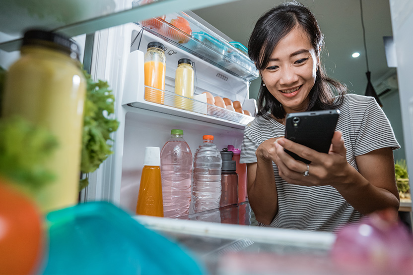 Young Asian woman looking at her smartphone with her refrigerator door open.