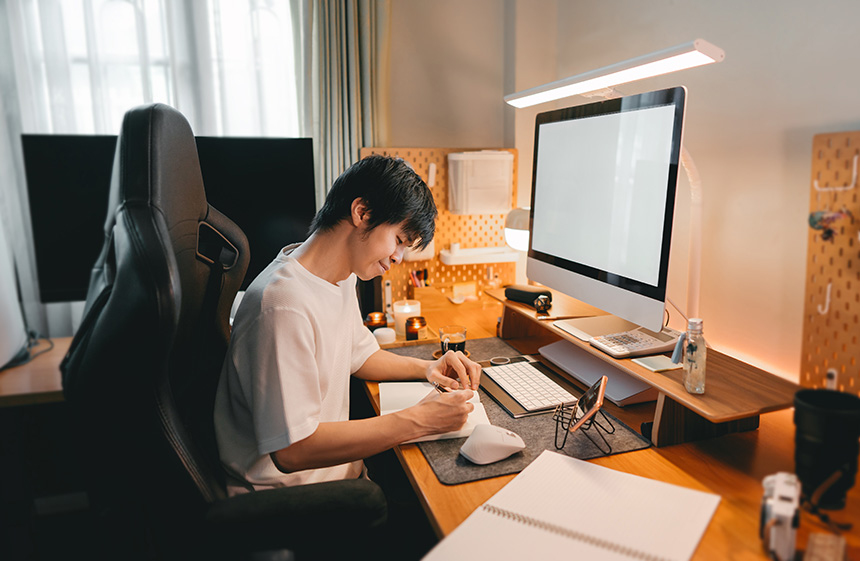 Person writing at a modern desk setup with computer and warm lighting.