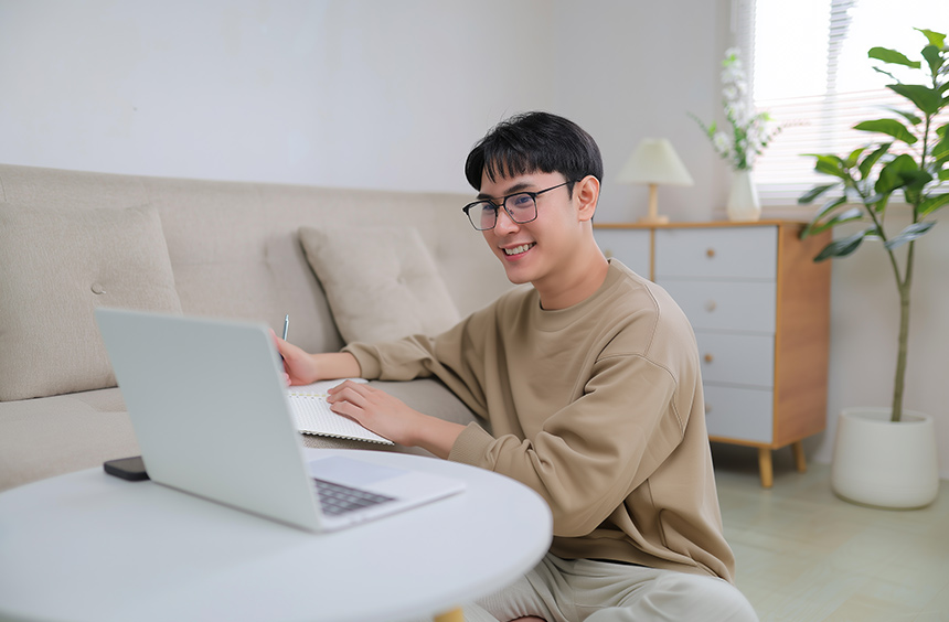 Young man taking notes while using a laptop at home.
