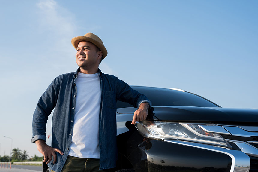 A man in a straw hat leans on a black car with a clear sky background.