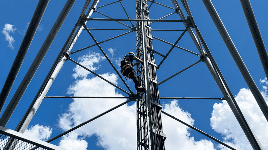 A Globe technician in full safety gear climbs the ladder of a tall metal cell tower against a blue sky, working to maintain essential connectivity infrastructure.