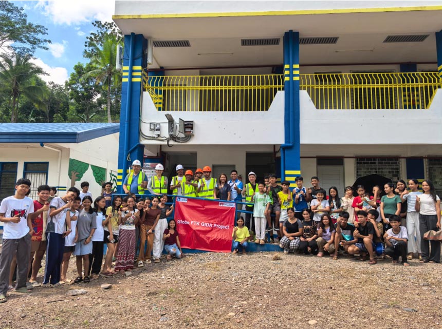Students, community members, and workers in safety gear posing together with a red Globe FTK GIDA Project banner in front of the two-story blue and white Sanghay National High School building.
