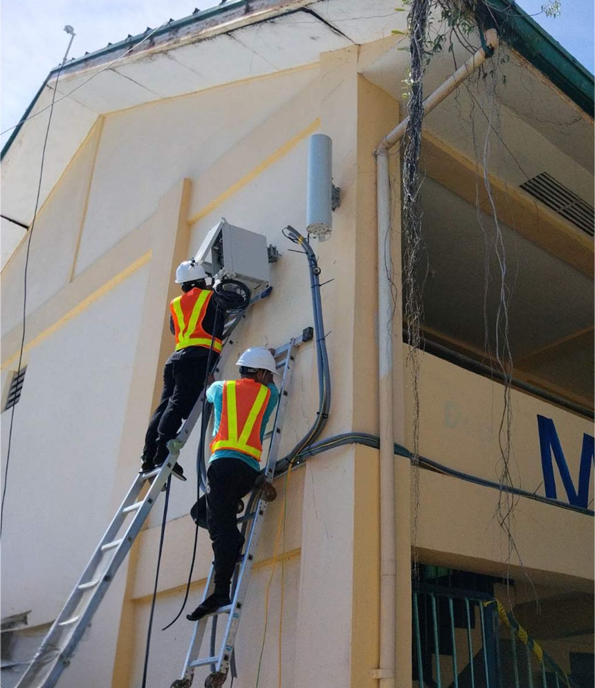 Two Globe technicians in safety gear working on a ladder to install internet equipment on a school building for the DICT Bayanihan SIM Project.