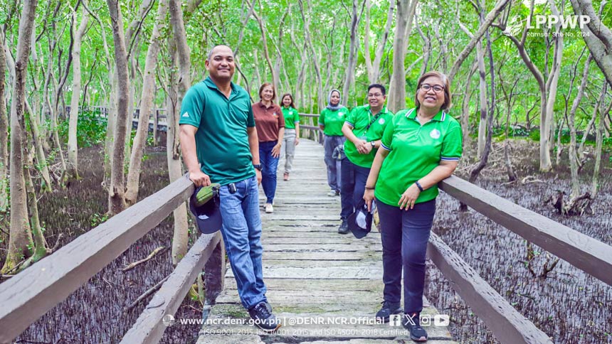 People in green shirts standing on a boardwalk in the middle of a forest.