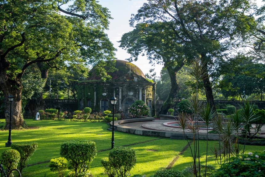 Old mossy chapel surrounded by grass, a fountain, and tall trees.