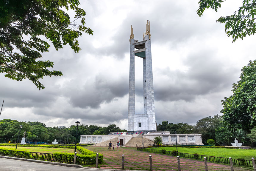 Quezon Memorial Shrine standing in the middle of grass and trees against a cloudy sky.