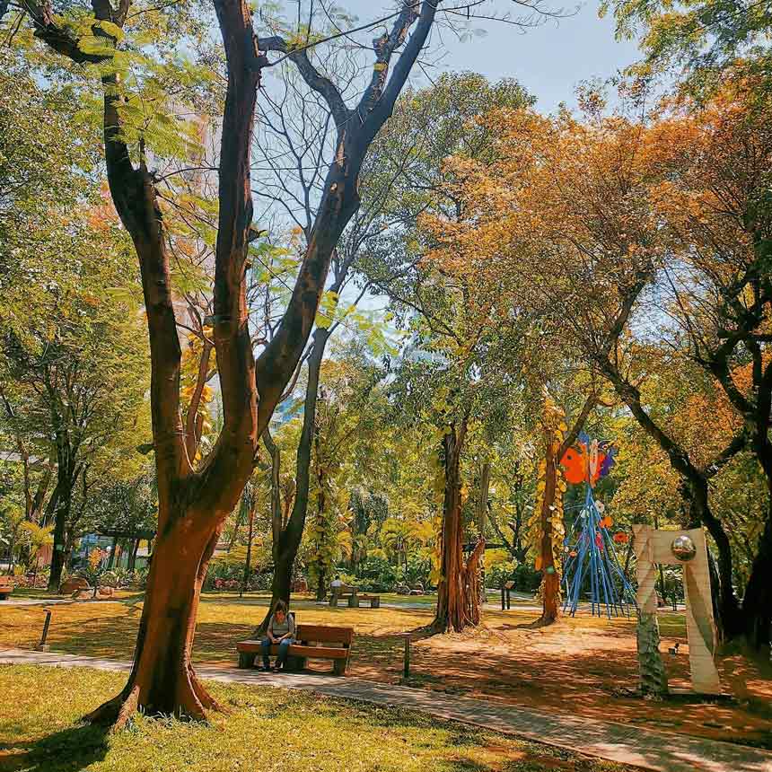 Small park with trees, benches, walkway, and sculpture.