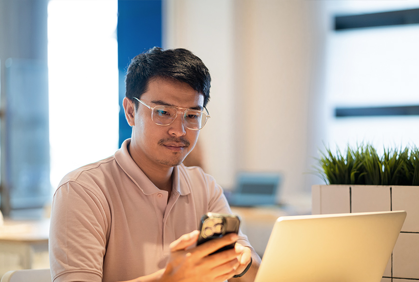 Asian male checks his mobile phone with laptop open