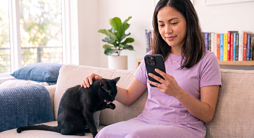 Woman sitting on a couch and petting her cat while using her phone.