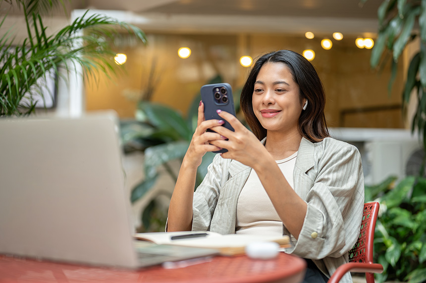 Woman using her smartphone while seated with a laptop.
