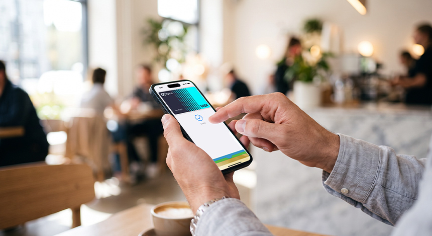 A customer sitting at a coffee shop table holding an iPhone to scan and pay for food using Apple Pay.