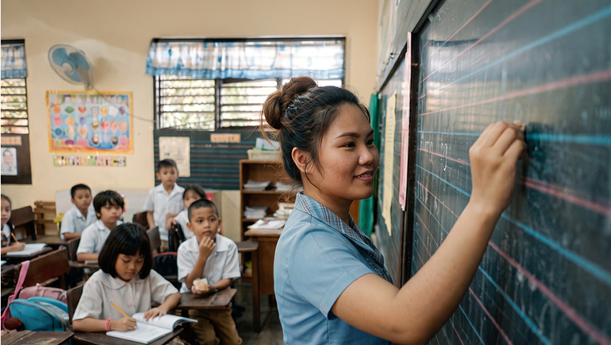 Teacher writing on a blackboard with chalk in a classroom full of young students sitting at desks.