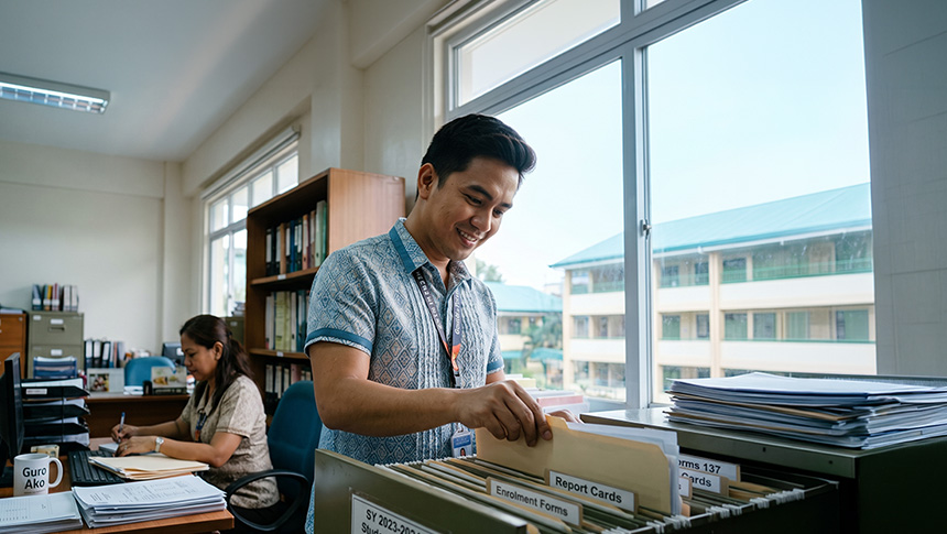 Teacher organizing student records in a filing cabinet while another teacher works at a desk.