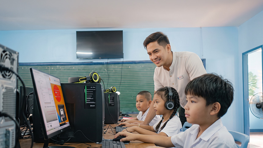 Teacher assisting young students as they use desktop computers in a computer lab.