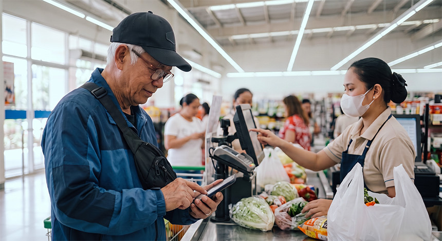 Senior man paying for groceries using a mobile phone at a supermarket checkout.