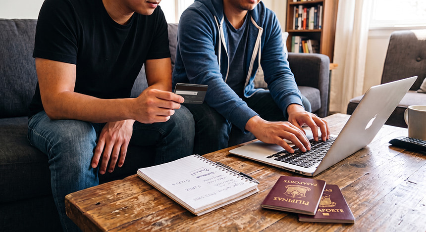 A couple sitting in front of a wooden table with a laptop and two Philippine passports on top, taking notes for an upcoming trip.